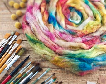May include: A collection of paintbrushes of various sizes and shapes, alongside a vibrant, multicolored skein of dyed wool roving. The wool displays a rainbow of colors, including pink, yellow, blue, and green, arranged in a spiral. The items rest on a wooden surface.