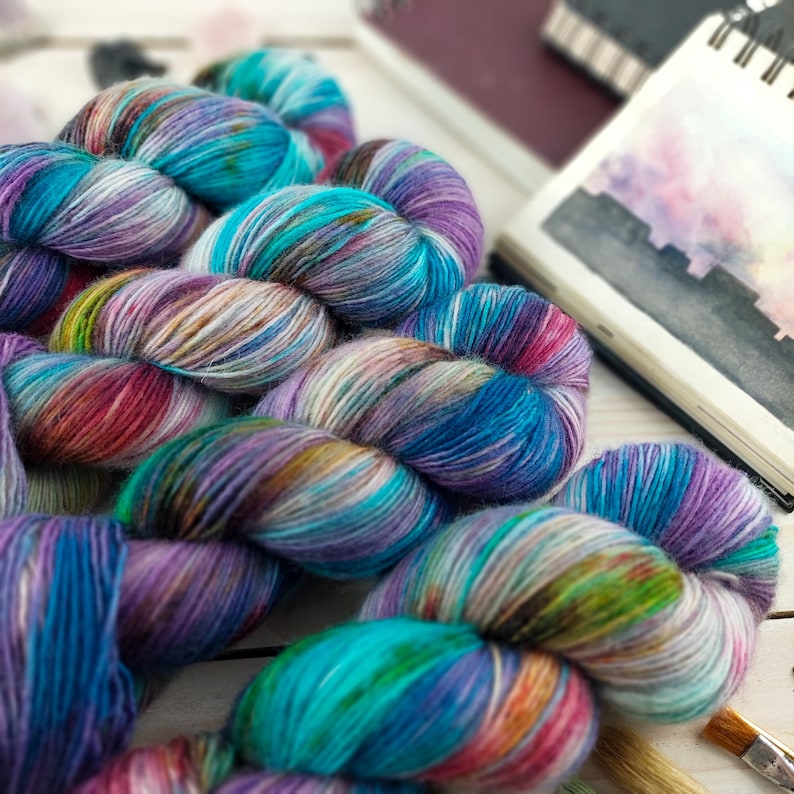 May include: Close-up of several skeins of colorful yarn in shades of blue, purple, green, and red. The yarn is arranged on a white wooden surface, with a sketchbook and paintbrushes in the background. The yarn appears to be hand-dyed.