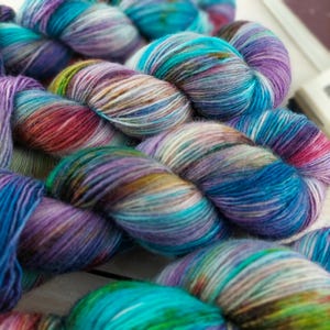 May include: Close-up of several skeins of colorful yarn. The yarn is a mix of blues, purples, greens, and reds, creating a vibrant, textured appearance. The yarn is likely for knitting or crocheting projects.