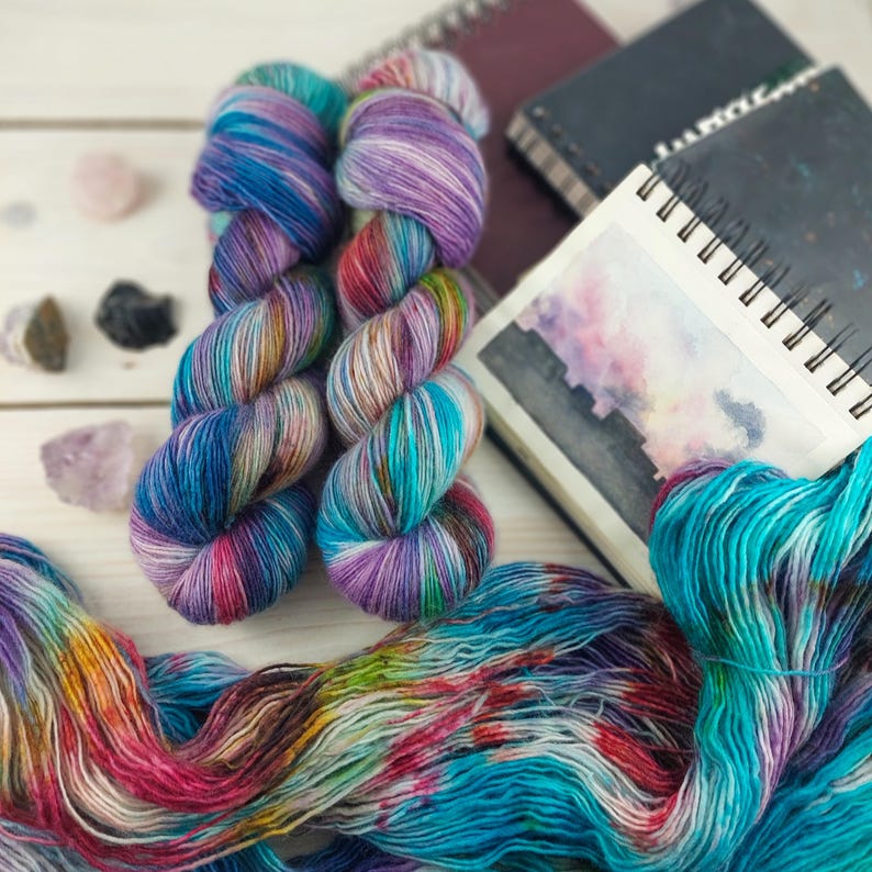 May include: Two skeins of colorful yarn in shades of blue, purple, and teal, with a watercolor painting of a cityscape and several small crystals. The yarn is arranged on a light-colored wooden surface.