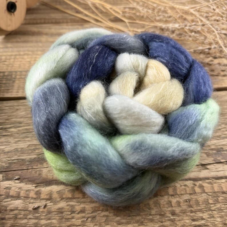 May include: A close-up of a braided yarn skein in shades of blue, green, and cream. The yarn is arranged in a circular pattern, with the colors blending together. The texture of the yarn is soft and fluffy, suggesting a fiber craft project.