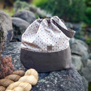 May include: A grey and cream drawstring bag with a floral pattern. The bag has a grey base and a cream top with a repeating pattern of small flowers in blue, pink, and green. A handle is attached to the top. Several skeins of yarn are in the foreground.