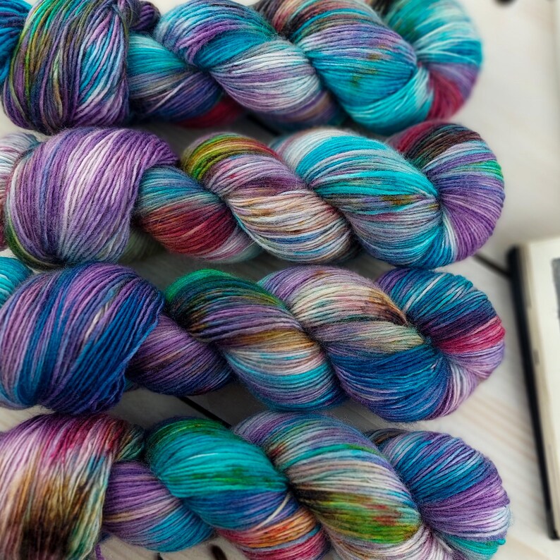 May include: Four skeins of hand-dyed yarn in a variety of colors. The yarn is a mix of blues, purples, greens, and pinks. The yarn is twisted into a rope-like shape. The yarn is on a light-colored wooden surface.
