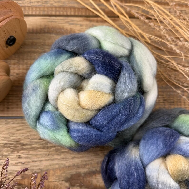 May include: A close-up of a braided fiber art piece in shades of blue, green, and cream. The fiber is arranged in a circular pattern, with a soft, textured appearance. The background is a wooden surface.