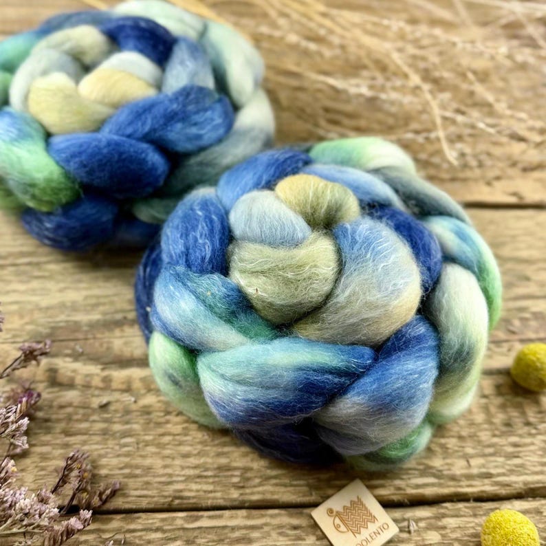 May include: Two hand-braided roving fiber bundles in shades of blue, green, and yellow. The wool is arranged in a circular pattern, with the colors blending together. The background is a wooden surface.