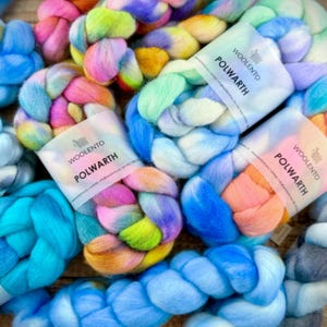 Può includere: Primo piano di lana merino intrecciata multicolore. L'immagine mostra matasse di lana nei toni del rosa, blu, giallo e verde. Ogni matassa ha un'etichetta con la scritta "WOOLENTO POLWARTH". La lana è disposta in un motivo visivamente accattivante.