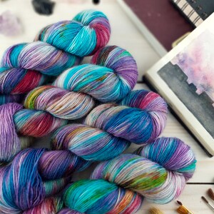 May include: Close-up of several skeins of colorful yarn in shades of blue, purple, pink, and green. The yarn is arranged on a white wooden surface, with art supplies and a sketchbook in the background. The yarn appears to be hand-dyed.