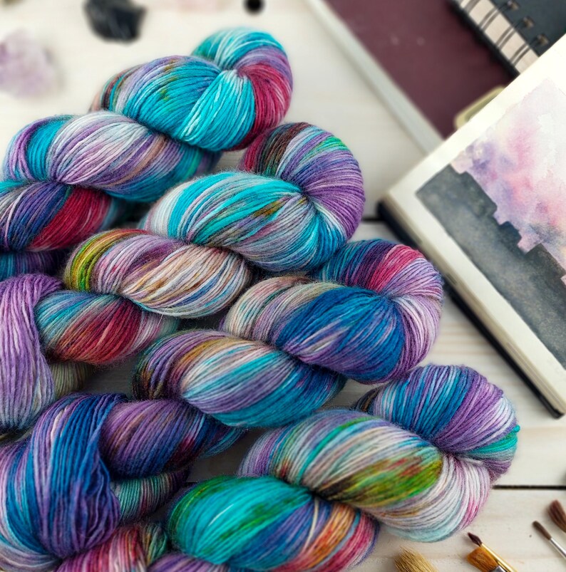 May include: Close-up of several skeins of colorful yarn in shades of blue, purple, pink, and green. The yarn is arranged on a white wooden surface, with art supplies and a sketchbook in the background. The yarn appears to be hand-dyed.
