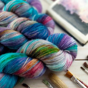 May include: Close-up of colorful yarn skeins in shades of blue, purple, green, and orange. The yarn is arranged on a white surface with paintbrushes and a blurred watercolor painting in the background. Ideal for knitting and crochet projects.
