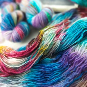 May include: Close-up of colorful yarn skeins and loose strands. The yarn features a vibrant mix of blues, purples, pinks, and yellows, creating a rainbow effect. The image showcases the texture and color variations of the yarn, ideal for knitting or crochet projects.