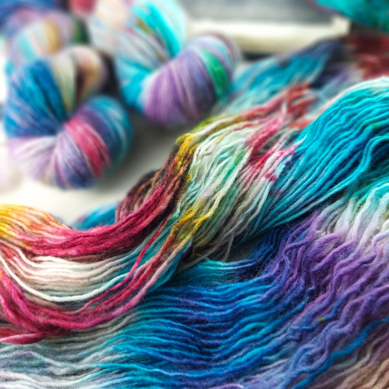 May include: Close-up of colorful yarn skeins and loose strands. The yarn features a vibrant mix of blues, purples, pinks, and yellows, creating a rainbow effect. The image showcases the texture and color variations of the yarn, ideal for knitting or crochet projects.