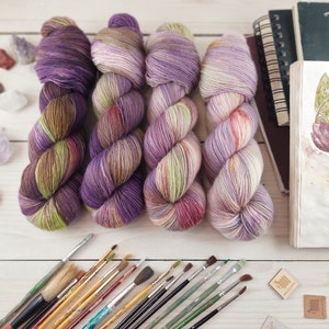 Könnte beinhalten: Vier Knäuel handgefärbter Wolle in den Farben Lila, Grün, Braun und Rosa. Die Wolle ist in einer Reihe auf einer weißen Holzoberfläche angeordnet. Es gibt Pinsel und ein kleines Holzschild mit der Aufschrift "Wool and Co.".