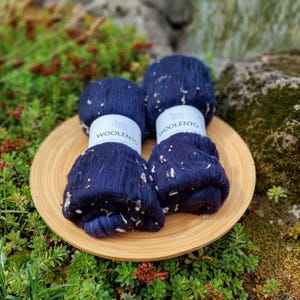 Può includere: Quattro gomitoli di lana blu navy con macchie bianche, etichettati "WOOLENTO", sono disposti su un piatto rotondo di legno marrone chiaro. La lana è circondata da fogliame verde e fiori rossi.
