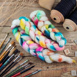 Può includere: Una spessa treccia di lana cardata multicolore con vivaci striature rosa, gialle, verde acqua e arancioni su base bianca, accompagnata da due rocchetti di legno con filo nero e una collezione di vari pennelli.