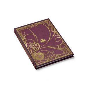May include: A burgundy journal with a gold embossed design featuring a clover and swirling patterns. The journal is closed and lying on a white surface.