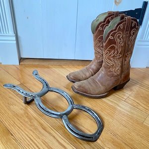May include: A metal horseshoe boot rack with two hooks. The rack is made of two horseshoes welded together. The rack is sitting on a wooden floor. Two brown cowboy boots are sitting next to the rack.