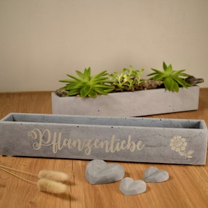 Pode incluir: Um vaso de concreto cinza com o texto "Pflanzenliebe" e um desenho de flor. O vaso está sobre uma superfície de madeira com três pequenas pedras cinzas em forma de coração.