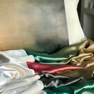 May include: A close-up of a bed with a white pillow and a multi-colored silk blanket. The blanket is made of different shades of green, gold, red, and white.