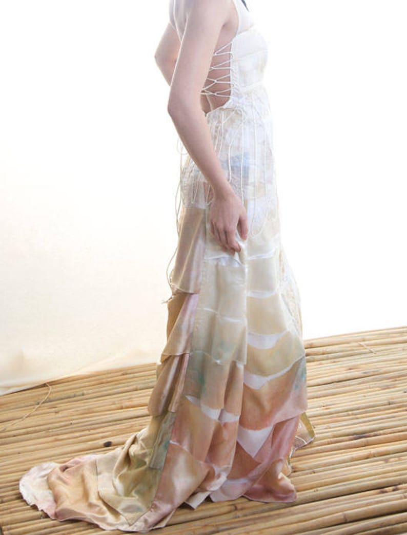 May include: A white, layered, and flowing gown with a lace-up back. The gown has a light pink and beige color gradient and is photographed on a bamboo surface.