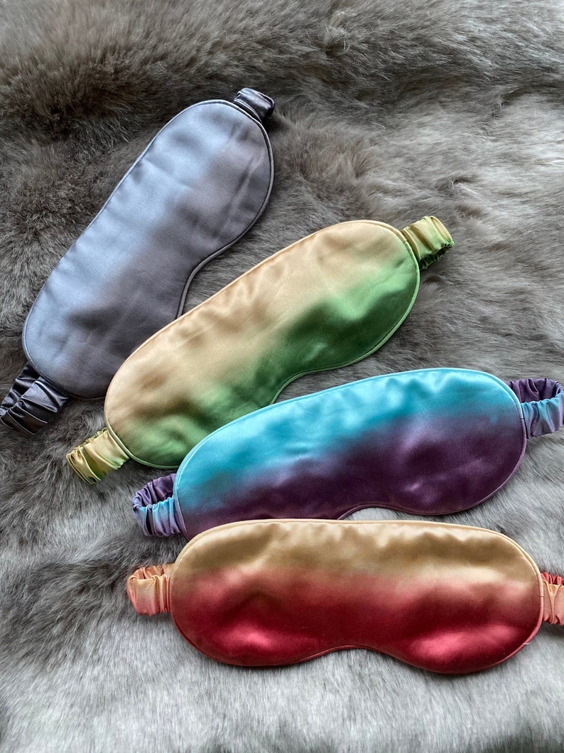 May include: Four silk sleep masks in different ombre colors. The masks are arranged in a row, with the top mask being a solid gray color, followed by a green and yellow ombre mask, a purple and blue ombre mask, and a red and orange ombre mask.