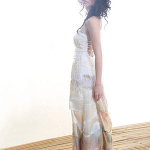 May include: A woman wearing a white, layered, and flowing dress with a lace-up back. The dress has a light pink and peach ombre effect at the bottom. The woman is standing on a wooden surface.