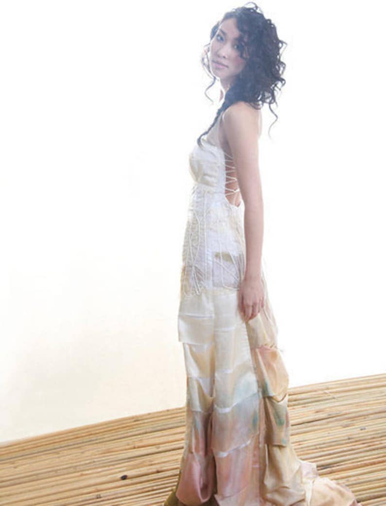 May include: A woman wearing a white, layered, and flowing dress with a lace-up back. The dress has a light pink and peach ombre effect at the bottom. The woman is standing on a wooden surface.