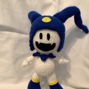 Peut inclure: Un jouet en peluche blanc crocheté avec un chapeau bleu et des accents jaunes. Le jouet a un large sourire aux dents et un chapeau de jester avec deux longues cornes recourbées.