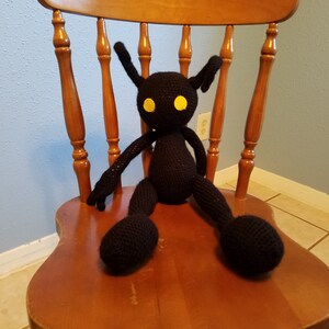 May include: A black crocheted plush toy shaped like a creature with large eyes and long arms and legs sitting on a wooden chair.