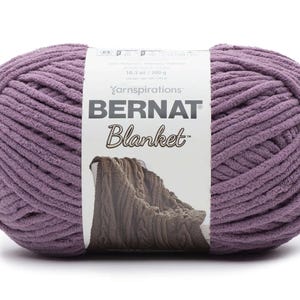 May include: A large, oval-shaped ball of Bernat Blanket yarn in a muted purple color. The yarn is thick and plush, with a soft, velvety texture. The label reads "BERNAT Blanket" and includes the weight: 10.5 oz / 300 g.