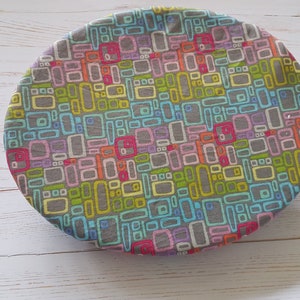 May include: A round, grey fabric bowl cover with a colourful geometric pattern of rectangles and squares in shades of blue, green, yellow, pink, and red.