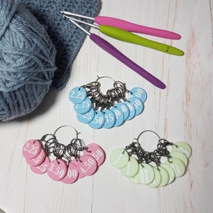 May include: An assortment of crochet tools and accessories. The image features a ball of blue yarn, a crocheted piece, three crochet hooks with pink, green, and purple handles, and stitch markers in blue, pink, and green.