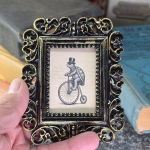 May include: Ornate black and gold picture frame with a whimsical illustration of a turtle riding a penny-farthing bicycle and wearing a top hat. The frame has a detailed, vintage design, perfect for displaying small artwork or photographs.