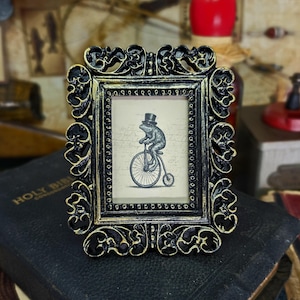 May include: Ornate black and gold picture frame with a whimsical illustration of a frog in a top hat riding a penny-farthing bicycle. The frame has a detailed, scrolling design and a vintage aesthetic. The artwork is set against a backdrop of handwritten script.