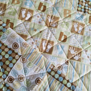May include: A baby quilt with a patchwork design featuring various fabrics with different patterns. The quilt is made up of squares with different colors and patterns, including brown, blue, green, and white. The quilt has a quilted texture and is made of soft fabric.