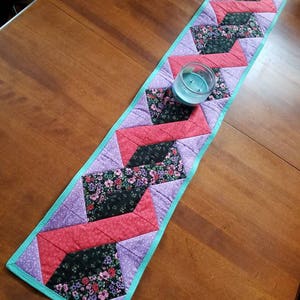 Op de afbeelding: Een turquoise, paarse, zwarte en rode gequilte tafelrunner met een zigzagpatroon. De runner ligt op een houten tafel met een turquoise kaarsenhouder.