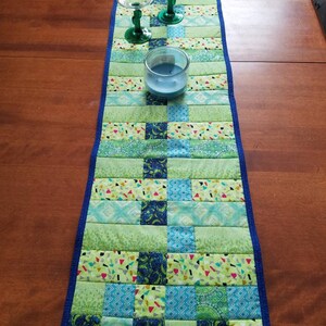 Può includere: Un runner da tavolo in patchwork verde e blu con un bordo blu. Tre calici di vetro verde e una candela blu sono sul runner.