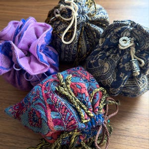May include: Four small fabric drawstring pouches with varied patterns and textures. One is solid purple and blue, while the others feature intricate woven designs in shades of navy, gold, red, and teal, accented with decorative metal beads and tassels.