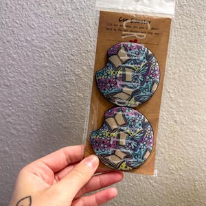 May include: Two car coasters with a colorful floral and butterfly design. The coasters are packaged together and feature the text "Car Coaster" and a short quote. The coasters are designed to fit in a car's cup holder.