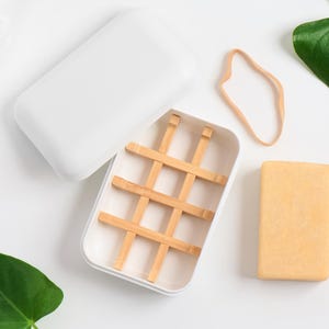 May include: White soap dish with a bamboo grid inside and a lid. A bar of soap is sitting next to the dish. There is a rubber band and green leaves in the background.