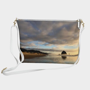 Può includere: Una borsa a tracolla in pelle bianca con chiusura a zip. La borsa presenta un'immagine stampata di una scena di spiaggia con una grande formazione rocciosa in lontananza, una spiaggia sabbiosa e un cielo nuvoloso.