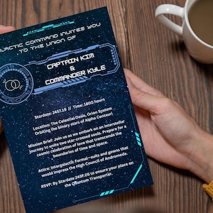 May include: A dark blue invitation card with white text, held by a person. The card's design features a space theme with the text "GALACTIC COMMAND INVITES YOU TO THE UNION OF CAPTAIN KIM & COMMANDER KYLE". A white mug with a brown liquid sits on a wooden surface.