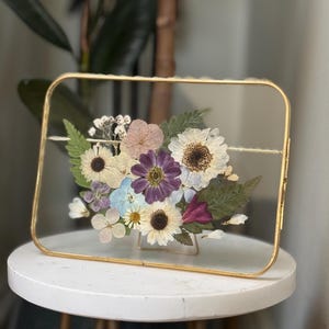 May include: A rectangular gold-framed glass display case filled with dried flowers. The arrangement includes white, purple, and yellow flowers, with green foliage. The case is resting on a small white table.