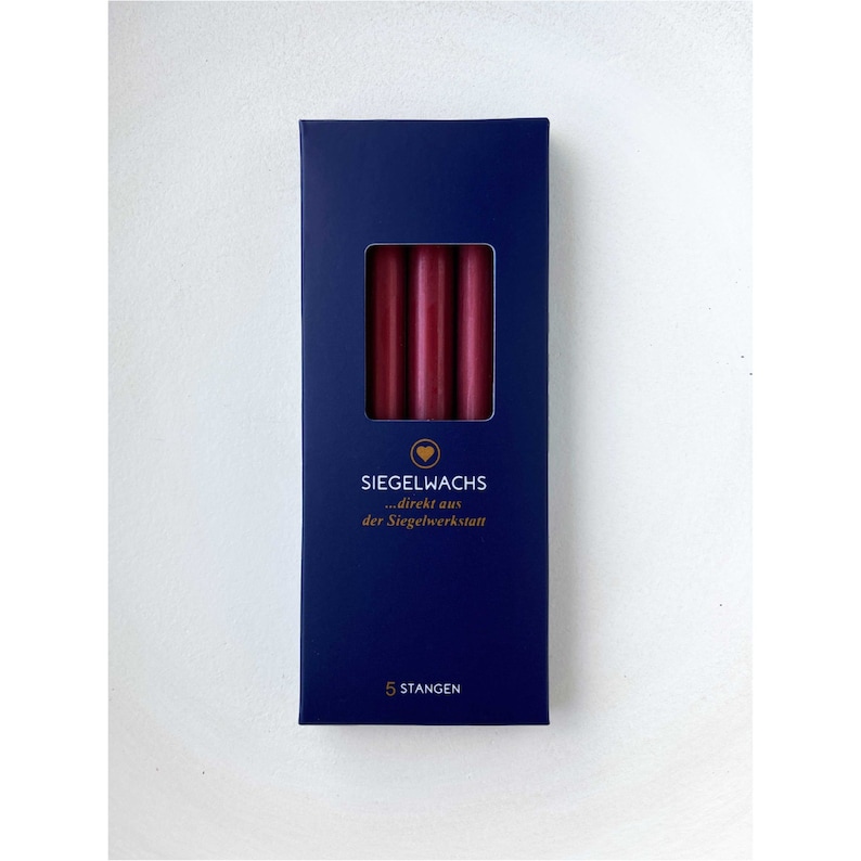 Puede incluir: Una caja de cinco barras de cera para sellar de color burdeos oscuro. La caja es azul marino con letras doradas que dicen "Siegelwachs...direkt aus der Siegelwerkstatt" y "5 Stangen".