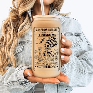 May include: A brown tumbler with a black and white graphic of a skeleton with flowers and the text "Some days I rock it. Some days it rocks me. Either way, we're rocking motherhood."