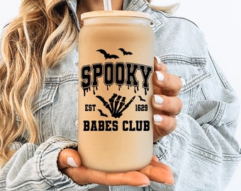 May include: A brown and black tumbler with the text "Spooky Babes Club" and a skeleton hand graphic. The tumbler has a straw and is being held by a person.