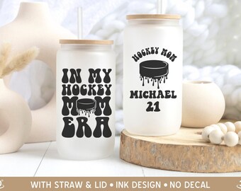 May include: Two frosted glass tumblers with lids and straws. One tumbler has the text "In My Hockey Mom Era" with a black hockey puck dripping black paint. The other tumbler has the text "Hockey Mom" with a black hockey puck dripping black paint and the text "Michael 21".