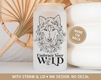 May include: White frosted glass tumbler with a black line drawing of a wolf's head with flowers around it. The text "Embrace Your WILD" is printed below the wolf. The tumbler has a bamboo lid and comes with a straw. The text "NAME ON BACK" is printed on a brown circle on the back of the tumbler.