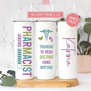 May include: Three white stainless steel tumblers with a black, green, and purple design. The tumblers are labeled "Pharmacist", "Training to Read Doctors' Handwriting", and "Katrina".