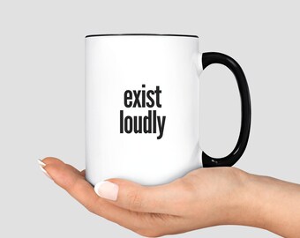 May include: White ceramic mug with a black interior and handle. The mug features the words "exist loudly" in a bold, black font. The mug is held in a person's hand against a light gray background.