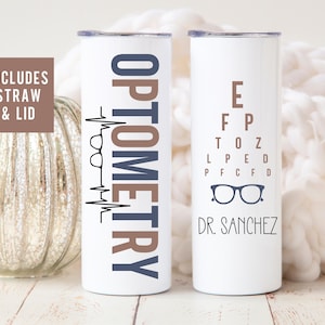 May include: Two white stainless steel tumblers with lids. One tumbler has the text "OPTOMETRY" in brown and blue lettering with a stylized heartbeat and glasses graphic. The other tumbler has an eye chart with the letters "E, FP, TOZ, LPED, PFCFD" and a pair of glasses graphic. The text "DR. SANCHEZ" is below the eye chart.